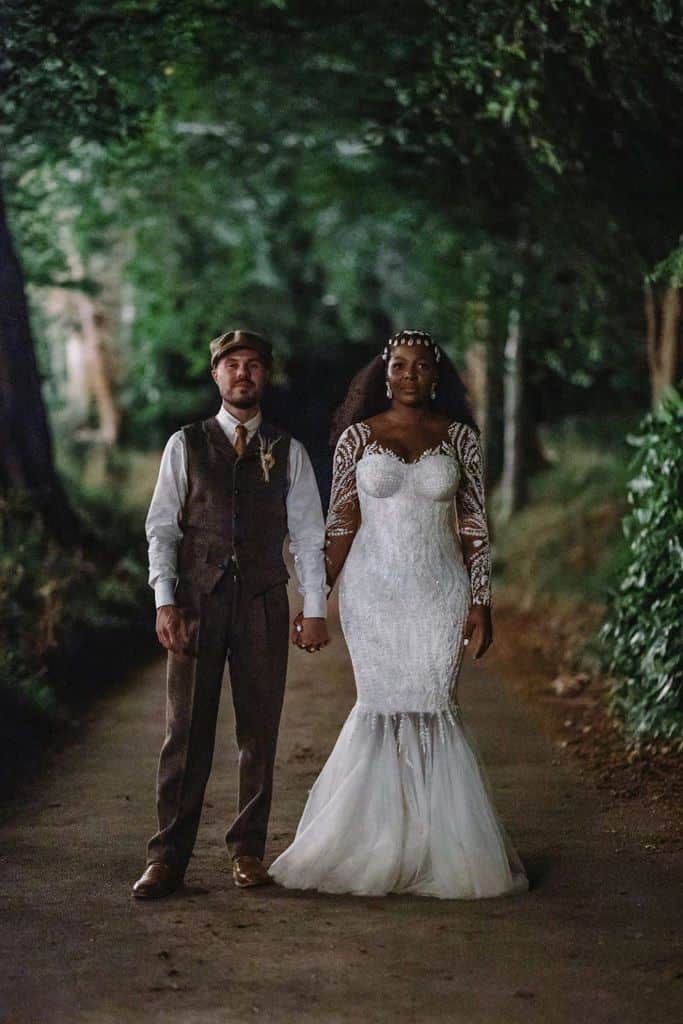 The couple are holding hands standing in the woods, the bride who has a dark skin tone is wearing natural makeup and bridal cowrie beads/shells.