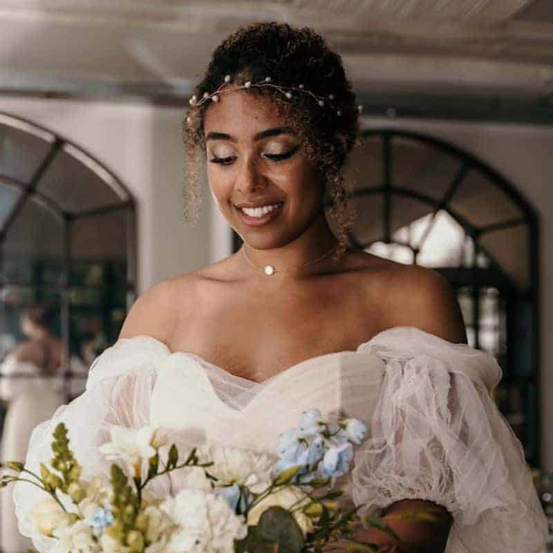 Mixed race bride smiling with her bouquet wearing her curly hair in an elegant updo accessorised with a pearl branch headband.