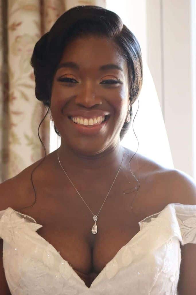 Black bride wearing a side parted bridal bun with hair wisps and soft glam natural makeup to accentuate her eyes.