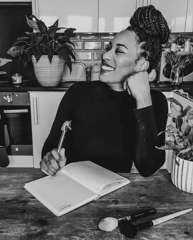 Aina.M, Afro Bridal Hair Academy Founder sitting at a table smiling with a notepad and pen in her hand.